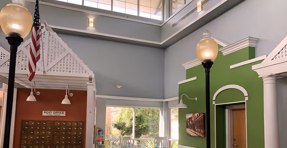 Interior common area with decorative storefront facades, a wall of mailboxes labeled 'Post Office', tall streetlamp-style lights and skylights above.