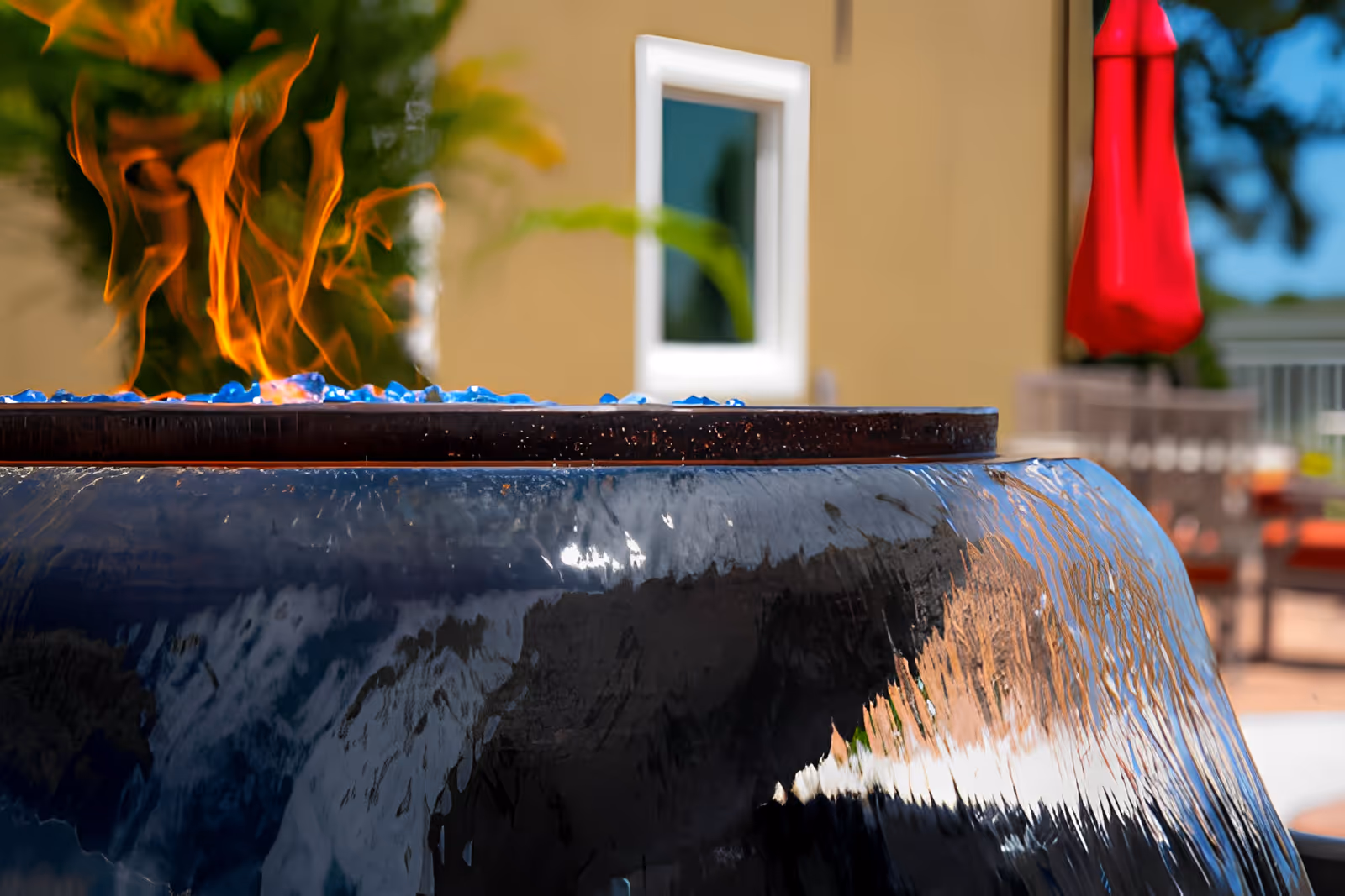 Close-up view of a modern outdoor fire pit with blue glass stones and visible flames, set in a patio area with a beige building wall, a white-framed window, a red patio umbrella, and outdoor furniture in the background.