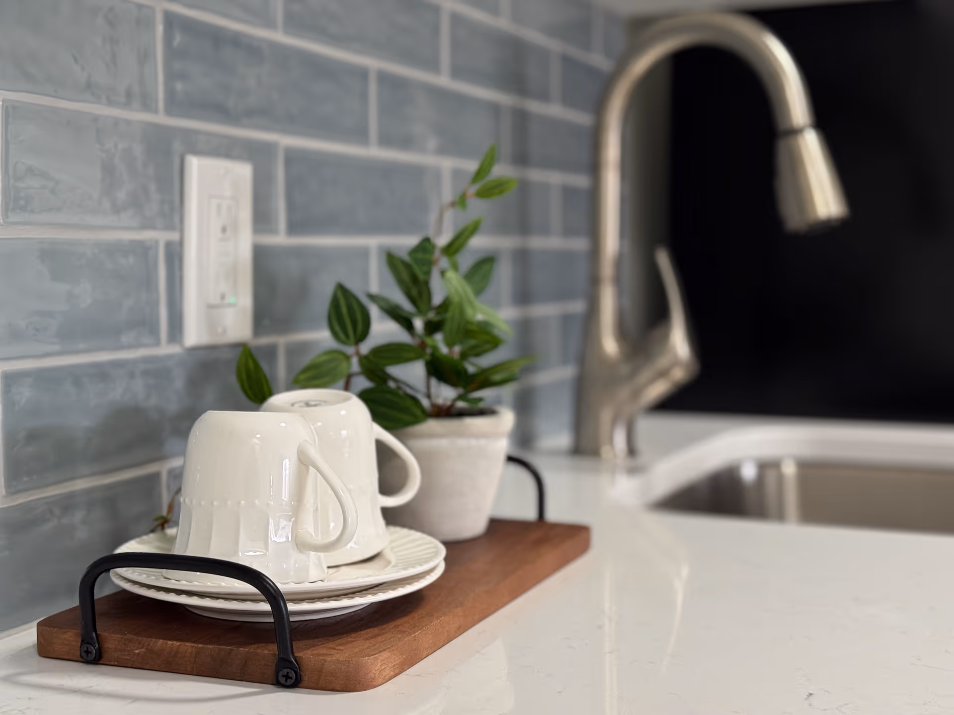 A kitchen countertop with a wooden tray holding white mugs and plates, a small potted plant, and a stainless steel faucet and sink against a gray tiled backsplash.