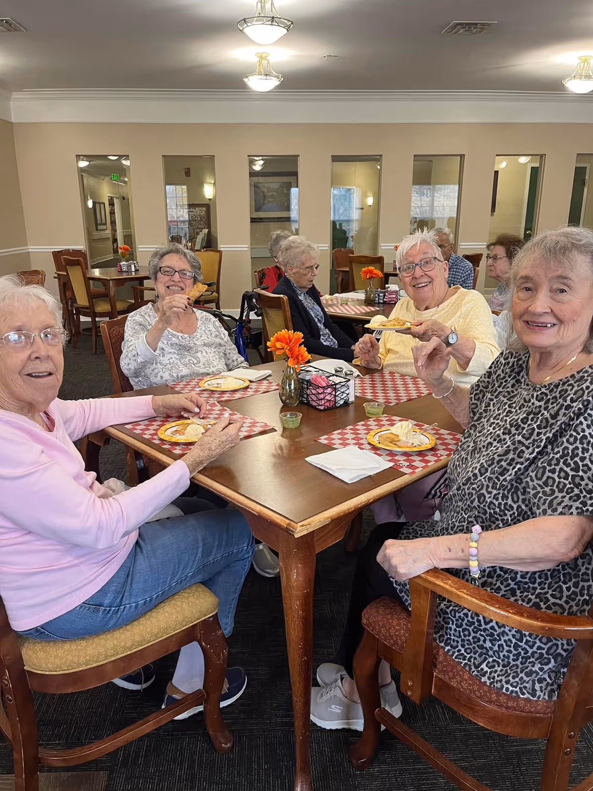 A group of elderly women sitting around a wooden dining table in a senior living facility, enjoying snacks and smiling. The table has red and white checkered placemats, small flower vases with orange flowers, and plates with food. The room has beige walls, mirrors, and additional tables and chairs in the background.
