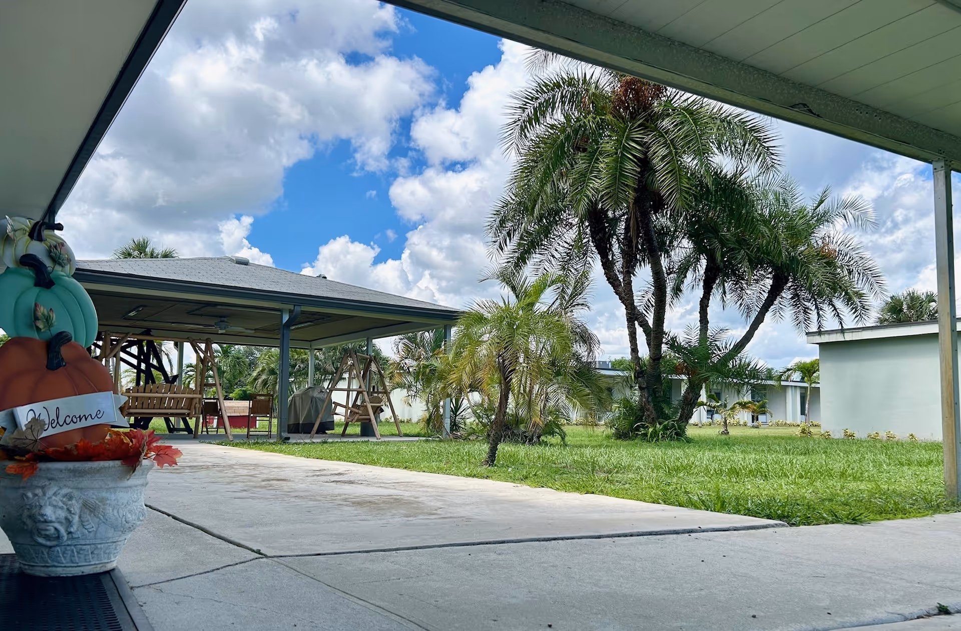 Outdoor view of a senior living facility with a covered patio area featuring wooden swings, a grassy lawn with palm trees, and a partly cloudy blue sky. A decorative planter with a pumpkin and a 'Welcome' sign is visible in the foreground.