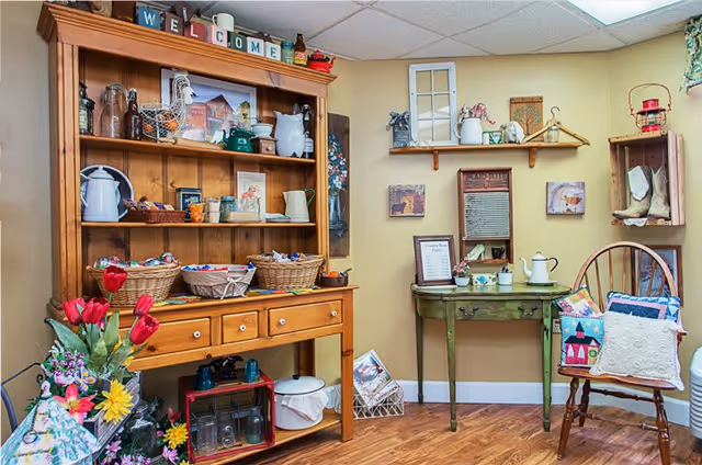 Decorative interior corner with a wooden hutch filled with baskets and ceramics, a small green table, and a chair with cushions.