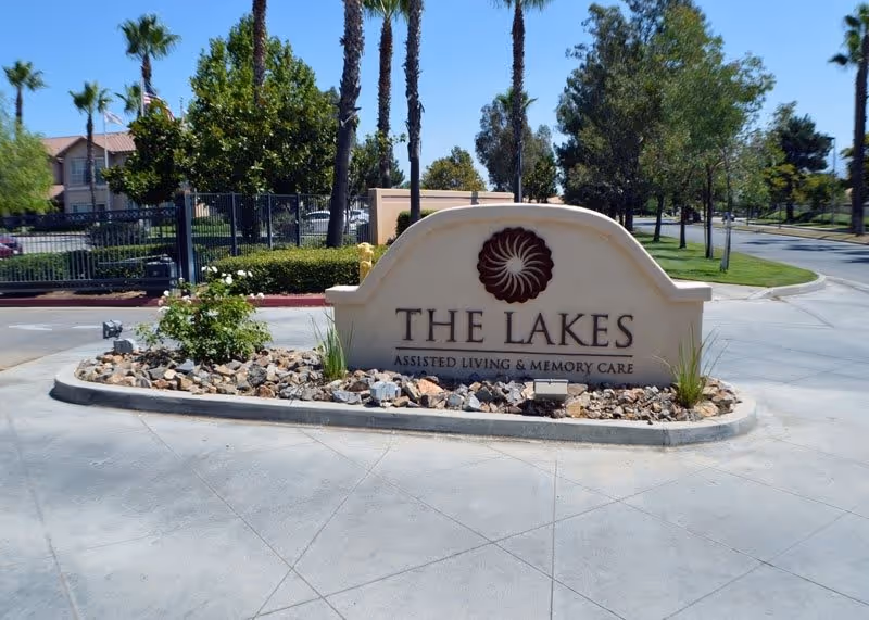 Entrance sign for The Lakes Assisted Living & Memory Care facility surrounded by landscaping with rocks and plants, palm trees and other greenery in the background along a paved driveway.