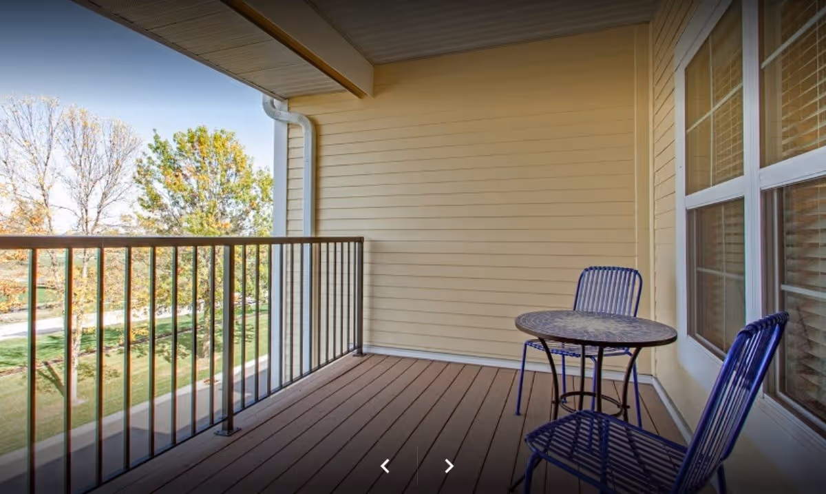 A covered outdoor balcony with a small round table and two metal chairs. The balcony has a railing and overlooks a green area with trees and a clear sky.