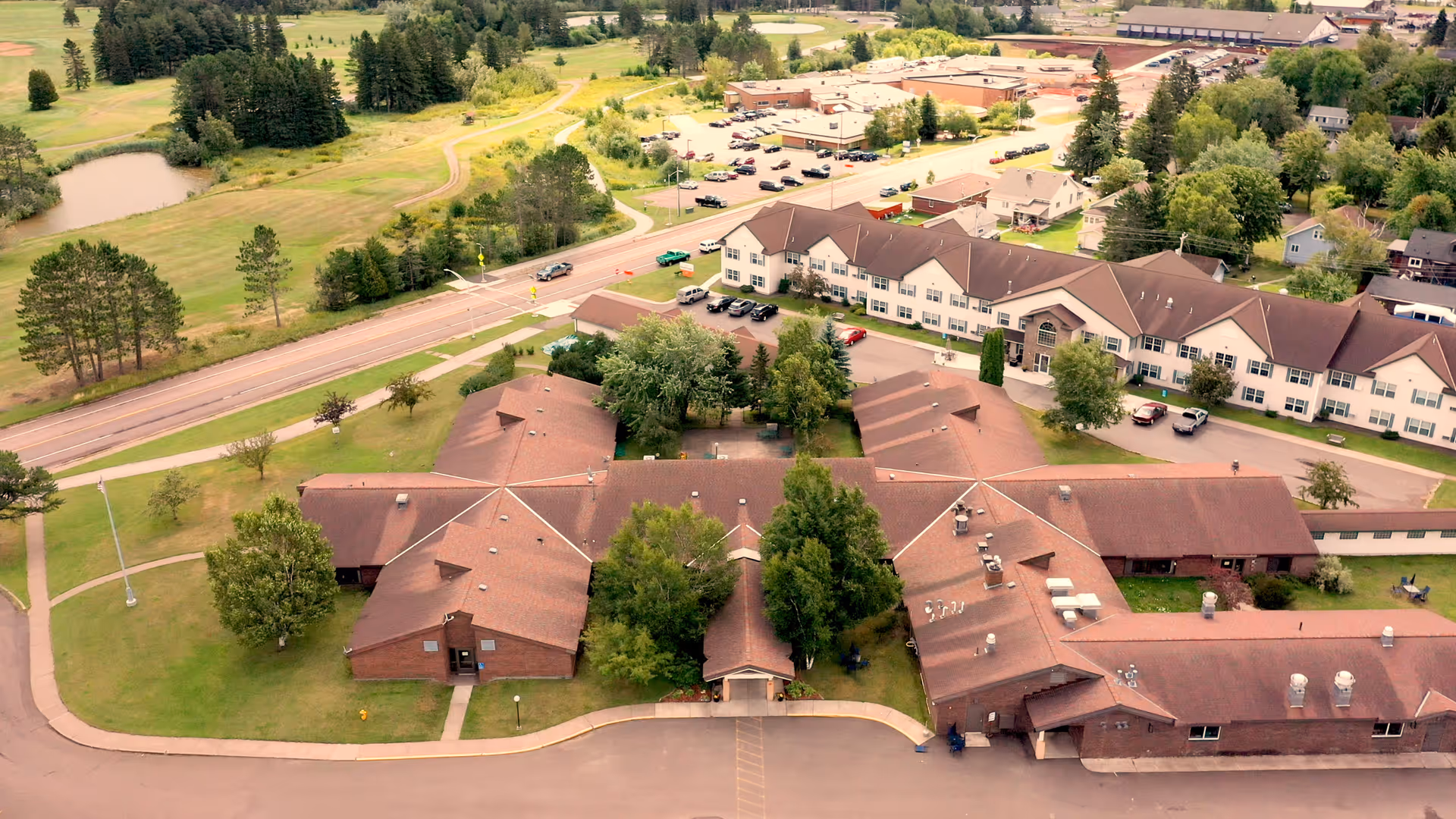 Aerial view of The Waterview Shores senior living facility showing multiple connected buildings with brown roofs surrounded by green lawns and trees. A road runs alongside the property with parked cars visible near the buildings. The surrounding area includes open grassy fields and scattered trees.