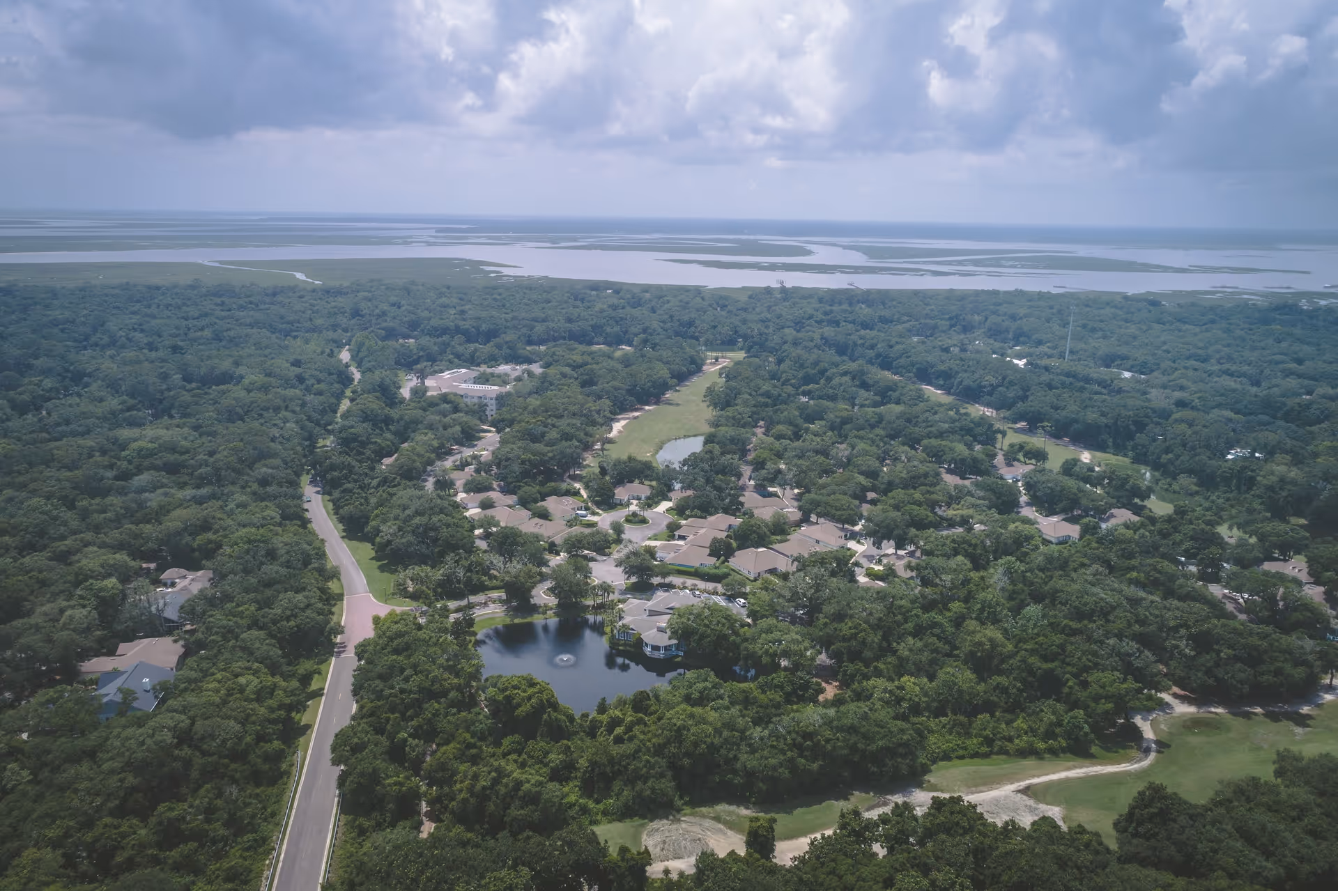 Aerial view of a residential community surrounded by dense green trees with a small pond featuring a fountain in the center. The community is near a large body of water under a cloudy sky.