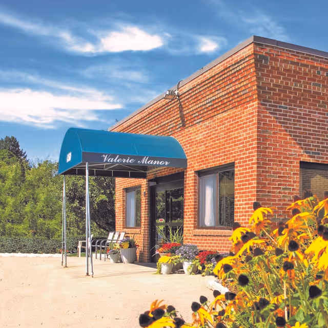 Exterior view of a brick building with a blue awning that reads 'Valerie Manor'. There are potted plants and flowers near the entrance, and yellow flowers in the foreground. The sky is partly cloudy.