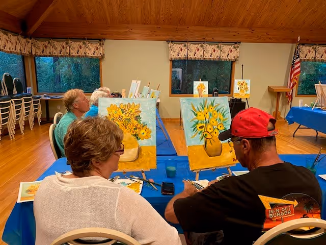 A group of people seated at tables in a room with wooden ceiling and large windows, engaged in painting yellow flowers on canvases set on easels. The room has blue tablecloths, and an American flag is visible in the background.