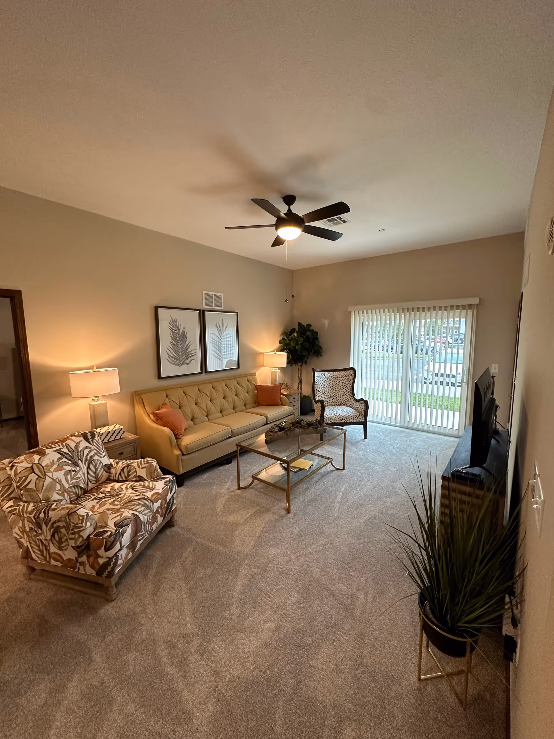 A cozy living room with a beige tufted sofa, two patterned armchairs, a glass coffee table, two side tables with lamps, a ceiling fan with light, a large potted plant, and vertical blinds covering a sliding glass door that leads outside.