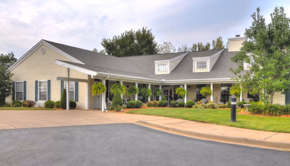Exterior view of a single-story senior living facility building with beige siding and a gray shingled roof. The building has multiple windows with dark shutters and a covered entrance supported by white columns. There are hanging green plants and well-maintained landscaping with bushes and trees surrounding the building. The driveway and a small lawn area are visible in the foreground under a partly cloudy sky.