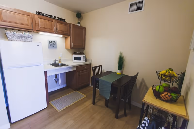A small kitchen and dining area with wooden cabinets, a white refrigerator, a microwave on the counter, a sink, and a dark wooden table with two chairs. The table has a green runner and a small plant. There is a two-tiered fruit basket with bananas, apples, and oranges on a side table. The wall above the cabinets has a decorative sign that reads 'HOME SWEET HOME'.