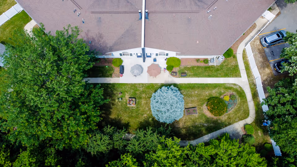 Aerial view of a senior living facility's outdoor garden area with a concrete walkway, two patio umbrellas, various trees, shrubs, and a parking area with cars visible on the right side.