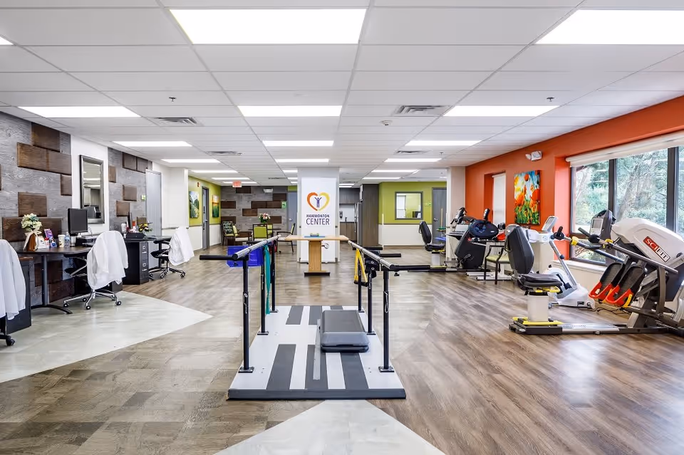 A spacious rehabilitation and exercise room at Hammonton Center featuring parallel bars for walking exercises in the center, several exercise machines including stationary bikes and a leg press on the right side near large windows, and desks with chairs and computers on the left side. The room has a mix of wood and tile flooring, bright overhead lighting, and colorful wall accents including a painting and a Hammonton Center logo on a central pillar.