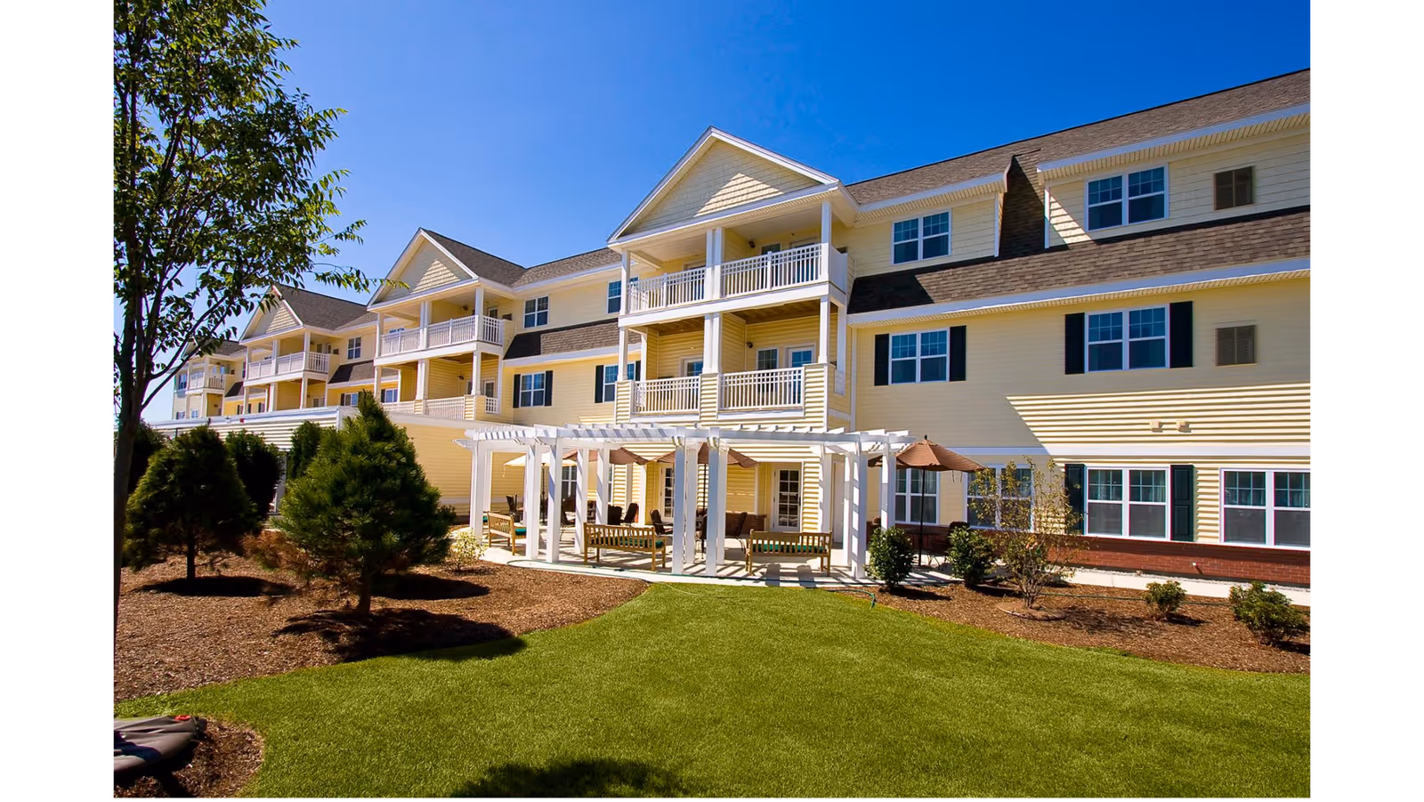 Exterior view of a multi-story senior living facility building with yellow siding and white balconies. In front of the building is a well-maintained lawn, landscaped bushes, small trees, and a white pergola with seating underneath. The sky is clear and blue.