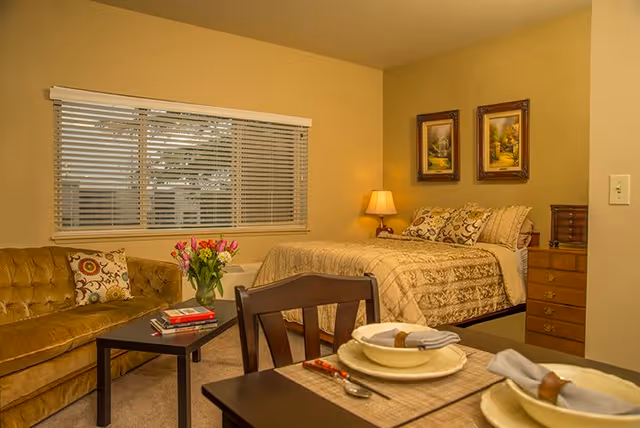 A cozy room with a bed covered in a beige patterned comforter, a wooden nightstand with a lamp, two framed paintings on the wall, a brown sofa with a decorative pillow, a coffee table with a vase of pink flowers and books, and a dining table set with plates, bowls, napkins, and cutlery. A window with blinds is visible behind the sofa.