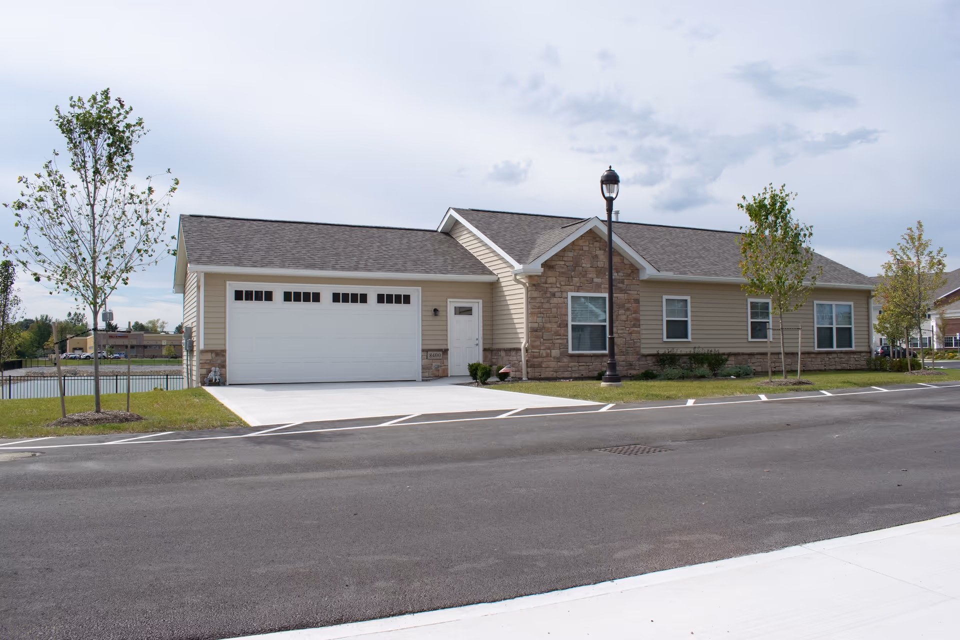 Exterior view of a single-story building with beige siding and stone accents, featuring a large white garage door, a small entrance door, several windows, a street lamp, and young trees planted along the sidewalk.