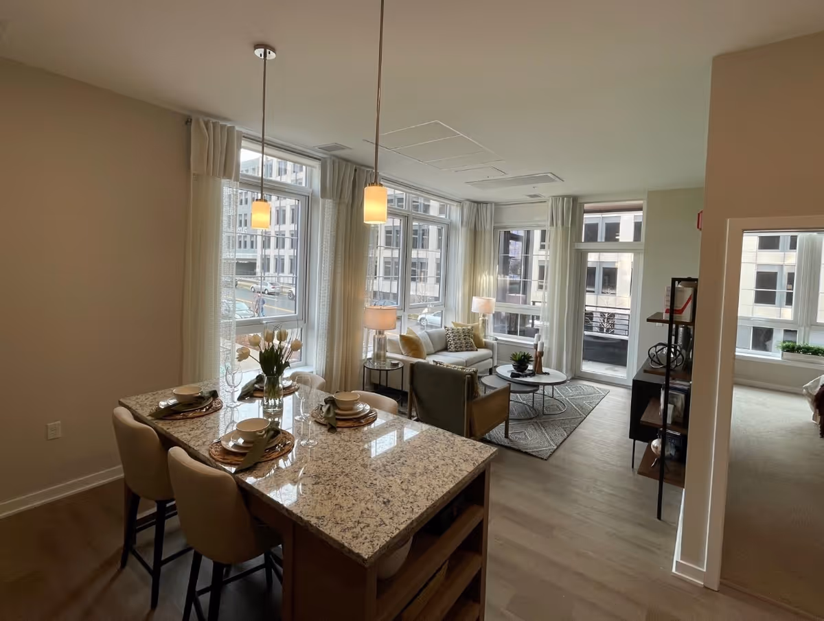 Open-plan apartment living area with a granite kitchen island set for four and a bright seating area by large windows.