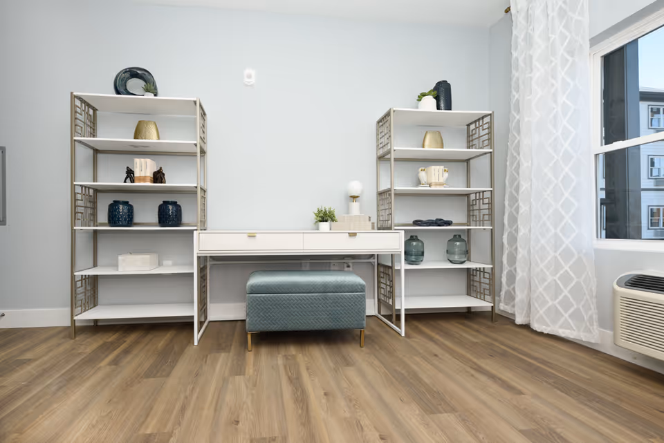 A modern interior room with light gray walls and wood flooring featuring two white and gold metal shelving units on either side of a white desk with two drawers. On the desk is a small plant and a lamp. In front of the desk is a blue cushioned bench. To the right is a window with white patterned curtains and an air conditioning unit below it.