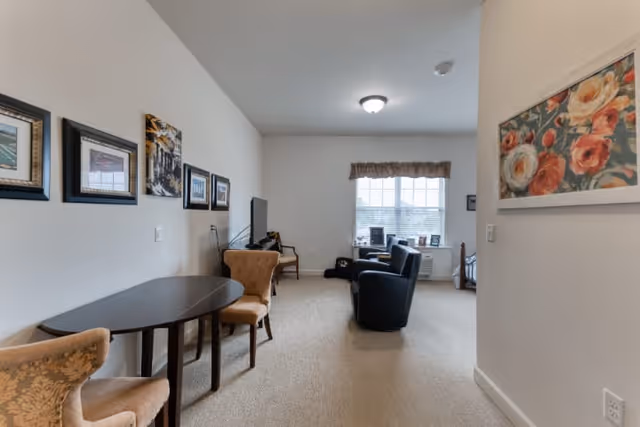 A cozy living room area with beige carpet and white walls. There is a dark wooden table with two upholstered chairs on the left side, and a TV on a stand against the far wall. Two black armchairs face the TV, positioned near a window with blinds and a valance. The walls are decorated with framed pictures and a floral painting.