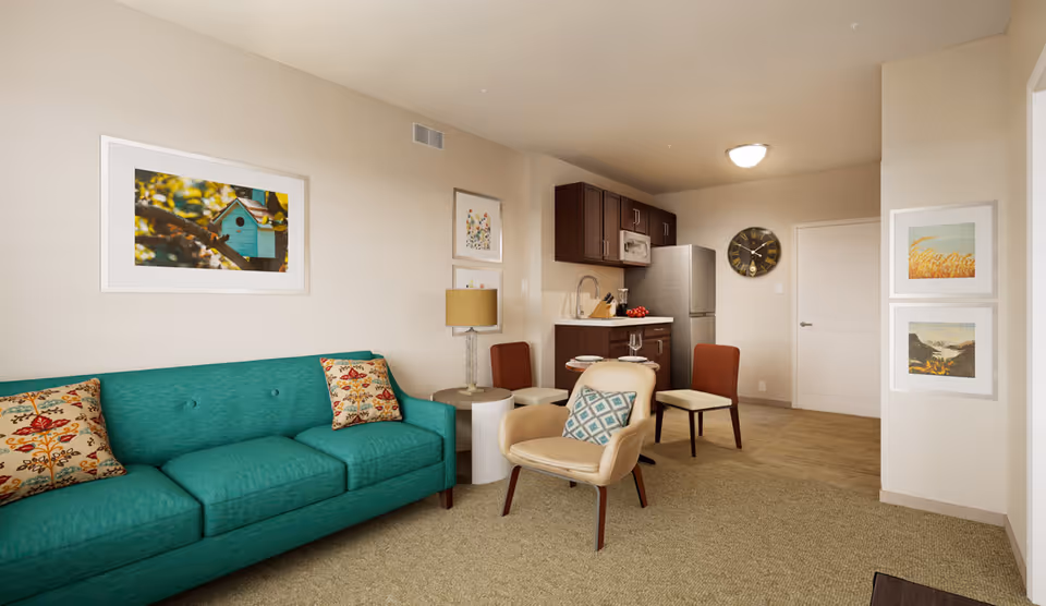 Bright living room with a teal sofa, armchairs, artwork on the walls, and a small kitchenette with a dining table in the background.