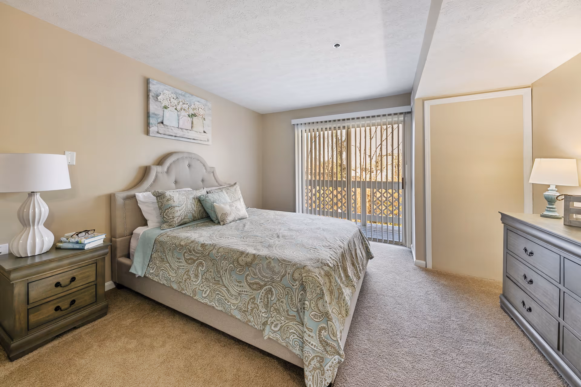 Well-lit bedroom with a bed, nightstands, dresser, and a sliding glass door with vertical blinds.