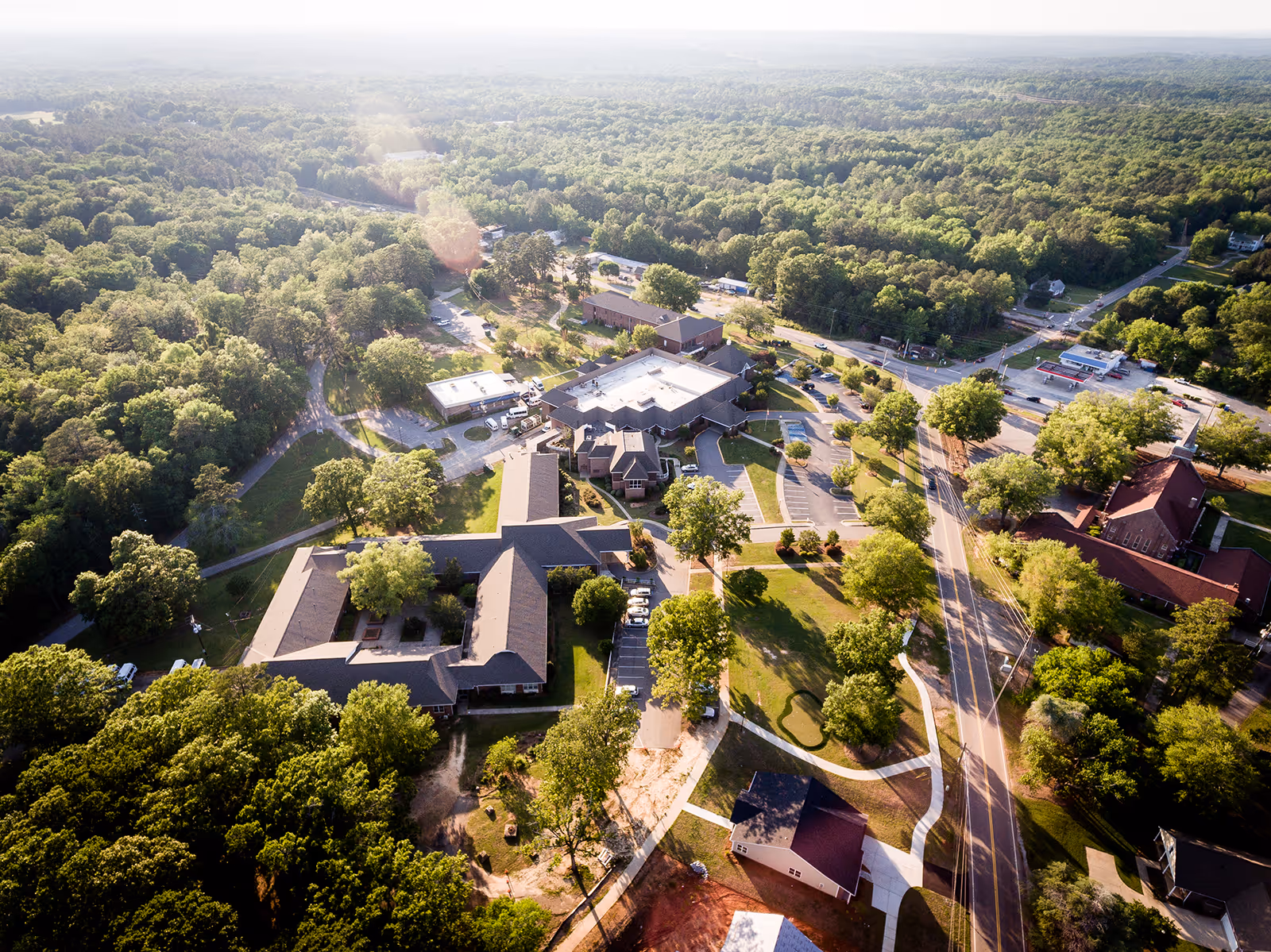 Aerial view of The Heritage At Lowman facility surrounded by dense green trees and roads, showing multiple buildings, parking areas, and pathways under bright daylight.