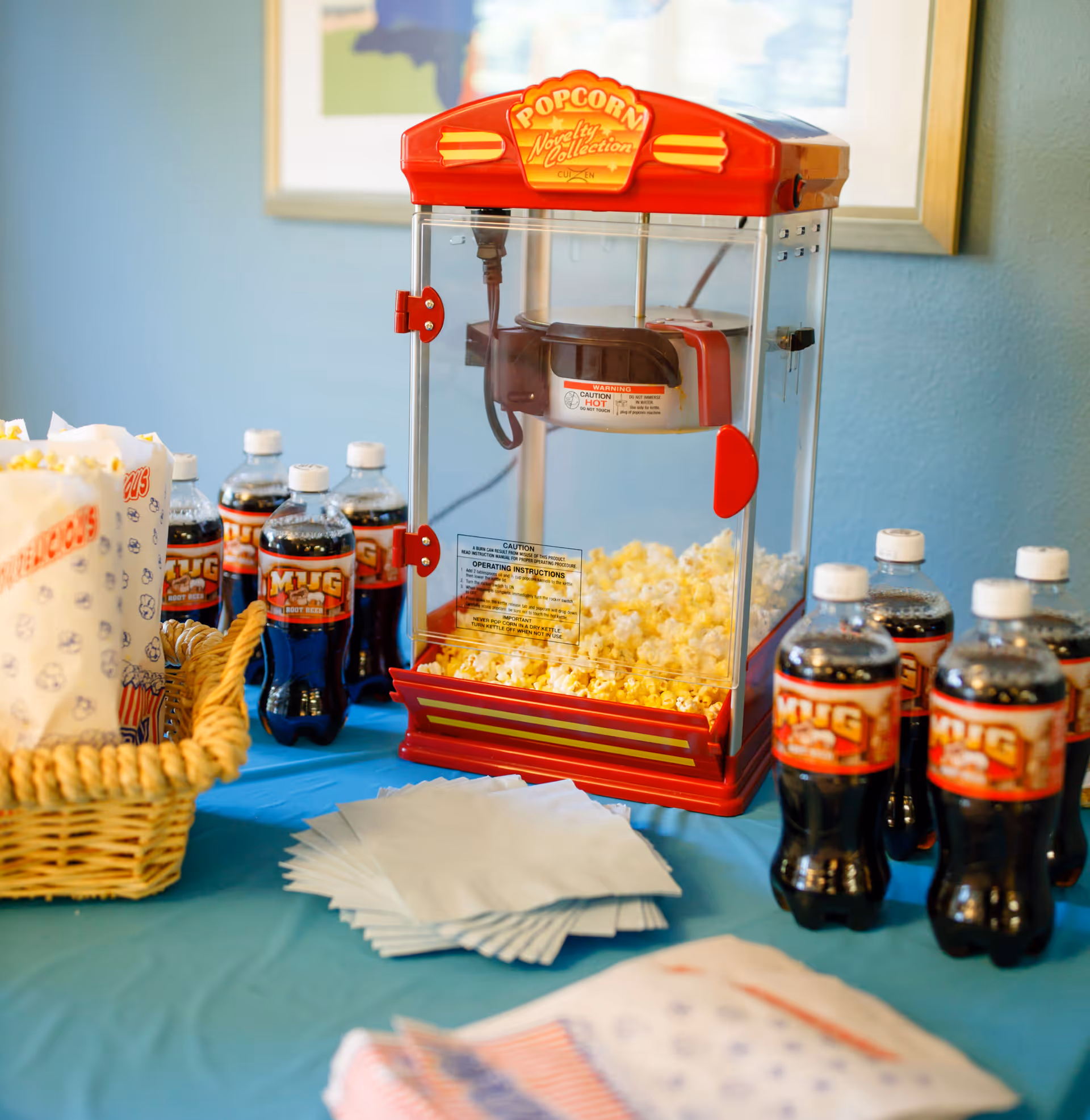 A red vintage-style popcorn machine filled with popcorn on a blue tablecloth. Surrounding the machine are several bottles of Mug Root Beer, a basket with popcorn bags, and a stack of white napkins. A framed picture is partially visible on the wall behind.