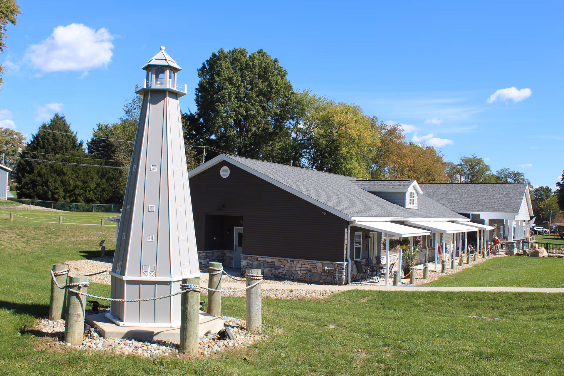 A single-story building with a gray roof and stone accents on the lower walls, surrounded by green grass and trees. In the foreground, there is a decorative lighthouse structure enclosed by wooden posts and rope. The sky is clear with a few clouds.