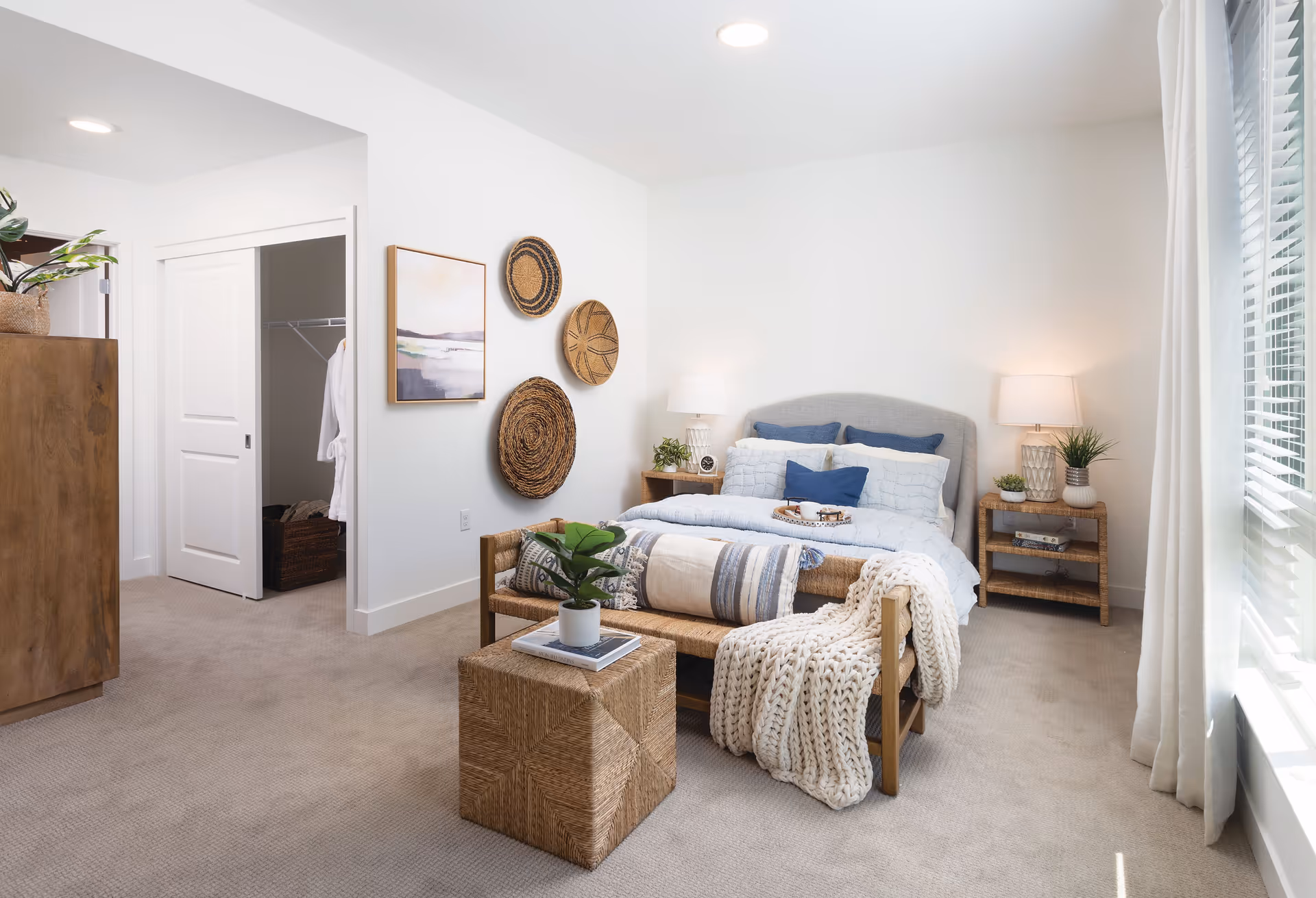 A bright and cozy bedroom with a neatly made bed featuring blue and white pillows and a light blue comforter. There is a wooden bench at the foot of the bed with a knitted throw blanket and decorative pillows. A small woven cube table with a potted plant and book sits in front of the bench. The room has beige carpeting, white walls, and a large window with white curtains letting in natural light. On the wall above the bench are three decorative woven baskets and a framed landscape painting. Two bedside tables with lamps and small plants flank the bed. An open closet with sliding doors is visible on the left side.