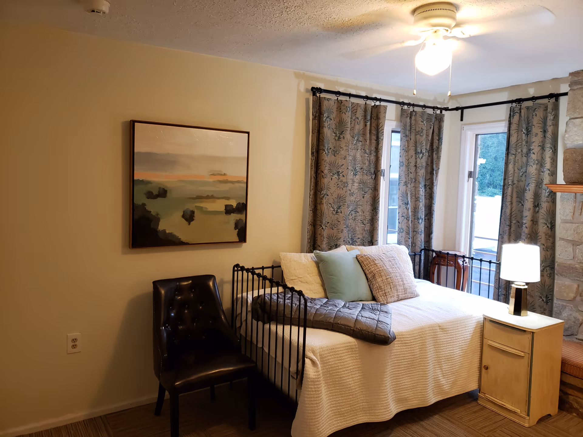 A cozy bedroom with a black metal bed frame, white bedding, and several pillows. There is a small wooden nightstand with a lamp on it next to the bed. A black leather chair is placed against the wall, which has a framed landscape painting. The room has windows with patterned curtains and a ceiling fan with a light. A stone fireplace is partially visible on the right side.