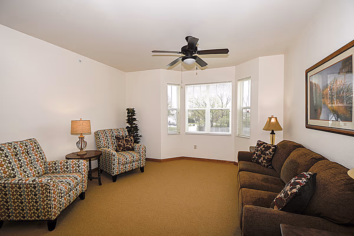 A living room with a brown sofa and two patterned armchairs. There are two table lamps on either side of the seating area, a ceiling fan with a light fixture, and a large bay window letting in natural light. A framed picture hangs on the wall above the sofa.