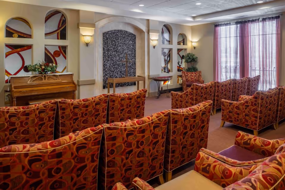 A small chapel-style gathering room with rows of patterned upholstered chairs facing an altar, a piano, and decorative wall niches beside large curtained windows.