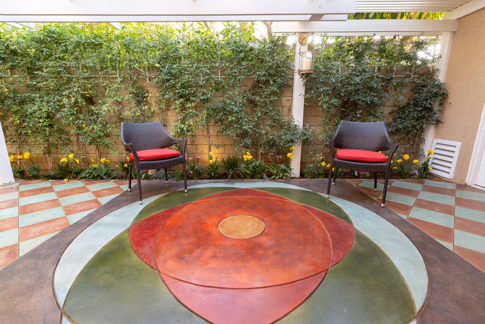Outdoor patio area with two dark wicker chairs with red cushions placed on a colorful circular patterned floor. The background features a wall covered with green climbing plants and yellow flowers along the base. A white pergola structure is overhead with a hanging lantern candle holder.