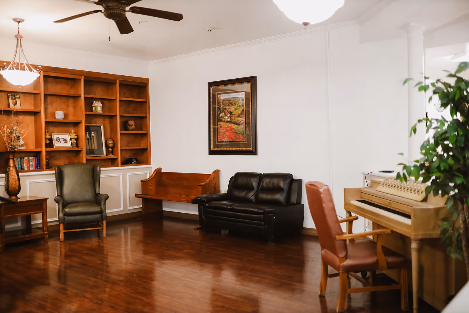 A cozy living room area with wooden flooring, a ceiling fan, and a chandelier. The room features a green armchair, a wooden bench, a black leather loveseat, and a wooden piano with a red cushioned chair. There is a large wooden bookshelf with various decorative items and books, and a framed landscape painting on the white wall. A green plant is visible near the piano.
