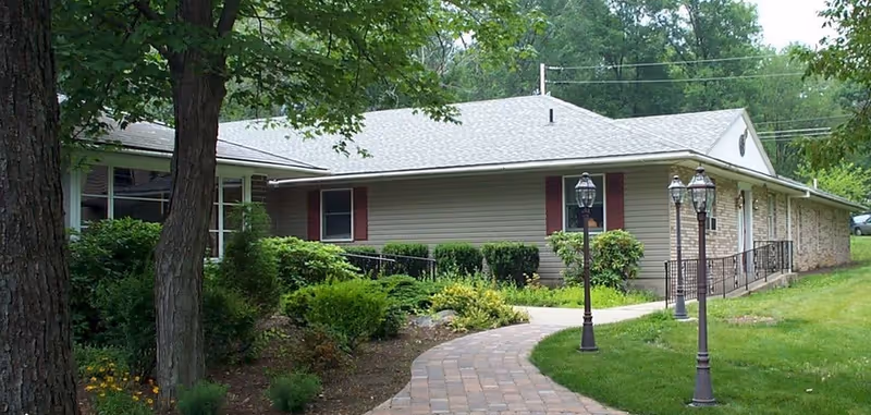 Single-story ranch-style building entrance with a curved brick walkway, lamp posts, and surrounding trees and shrubs.