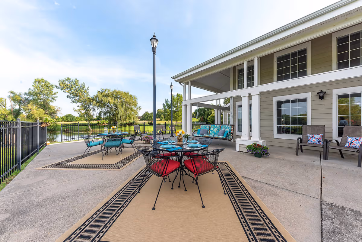 Outdoor patio area at Fort Scott Presbyterian Village with metal tables and chairs, some with red cushions and others with blue cushions. The patio features decorative outdoor rugs, a swing with blue cushions, potted plants, and a black metal fence overlooking a pond and green trees under a blue sky.