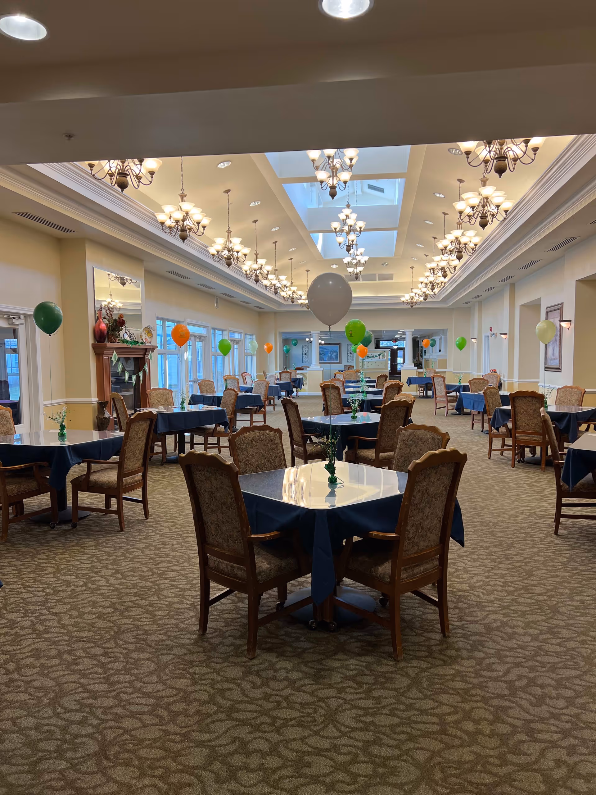 A spacious dining room with multiple tables covered in dark blue tablecloths, each surrounded by four upholstered chairs. The room is decorated with colorful balloons tied to the tables and has elegant chandeliers hanging from the ceiling with skylights. There is a fireplace on the left side and large windows along the left wall allowing natural light to enter.