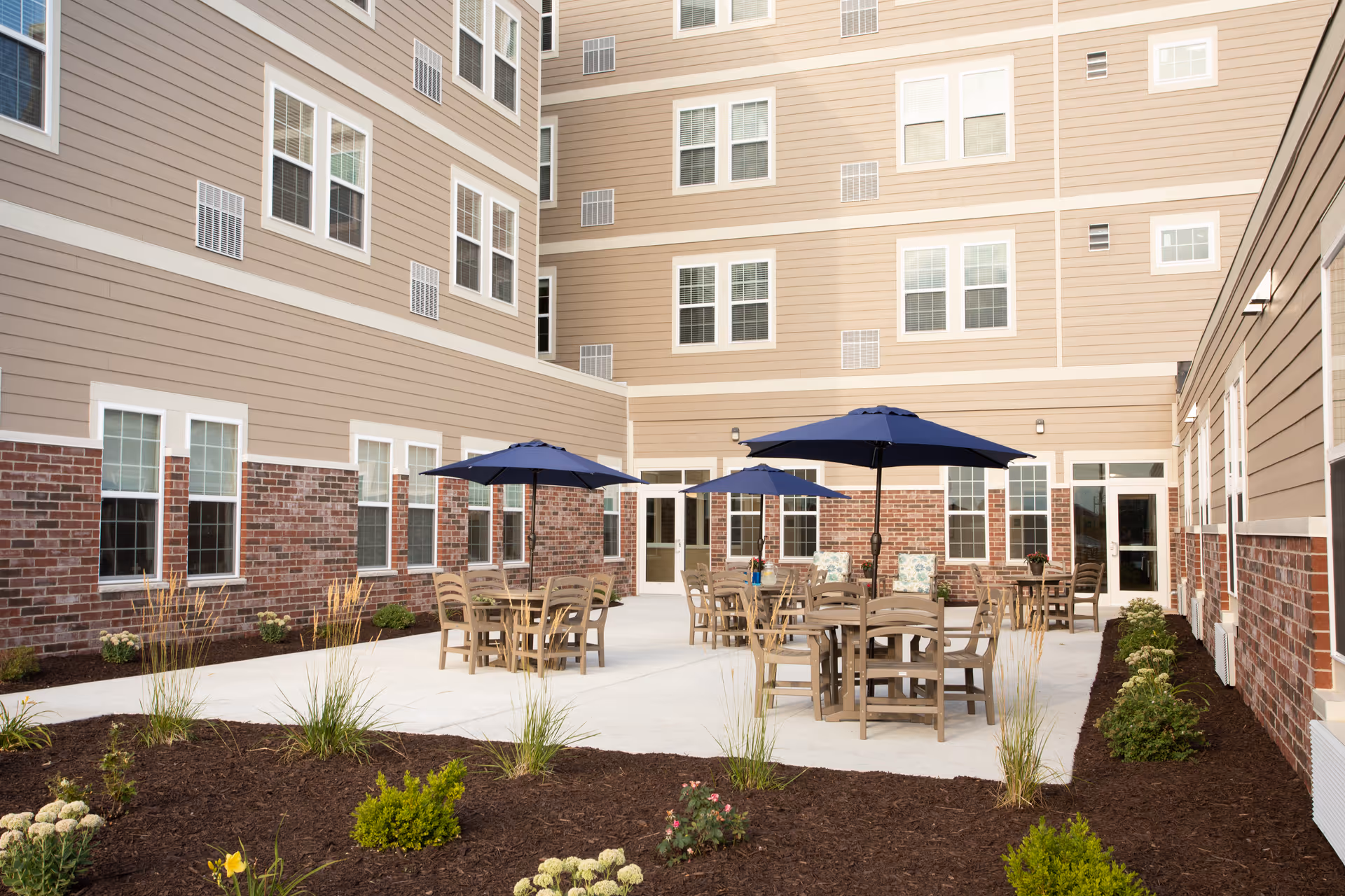 Outdoor courtyard area of a senior living facility with multiple round tables and chairs under blue umbrellas, surrounded by a beige and brick building with many windows and landscaped garden beds.