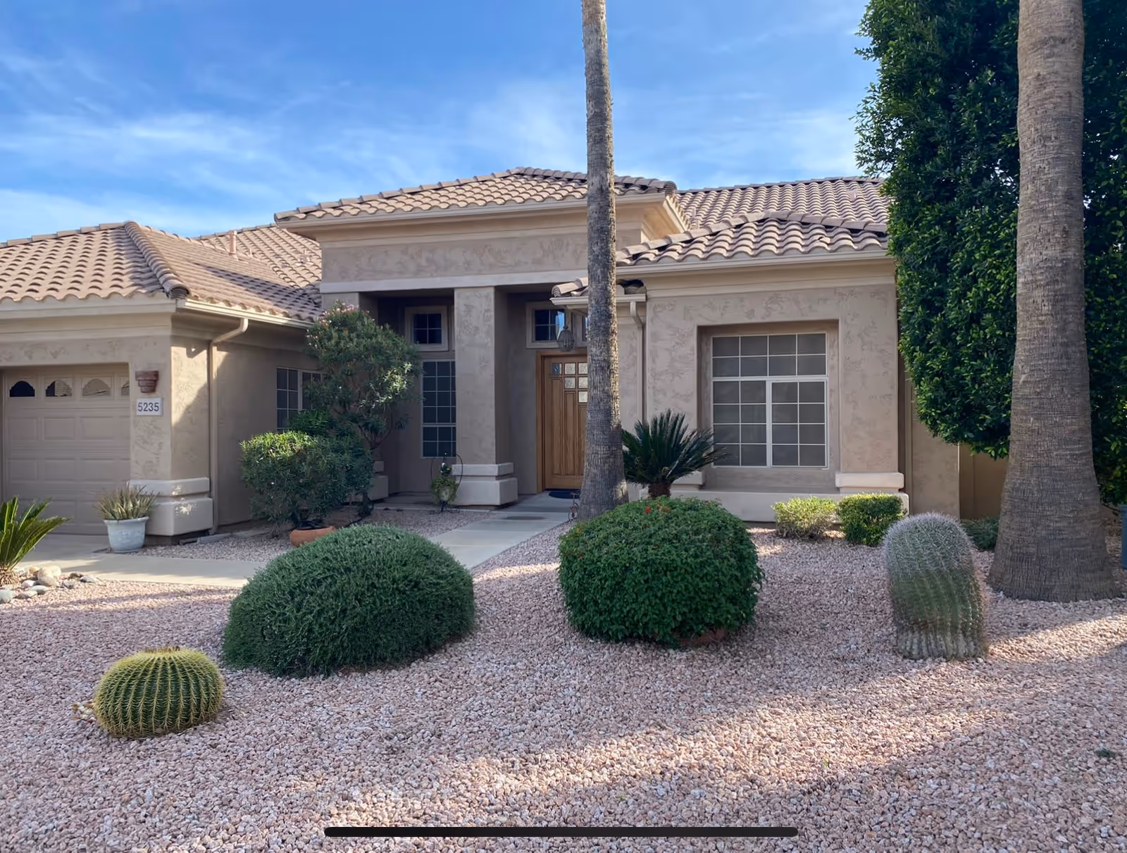 Front exterior view of a single-story house with a tiled roof, beige stucco walls, a wooden front door, and several windows. The front yard features desert landscaping with gravel, various bushes, cacti, and two tall palm trees.