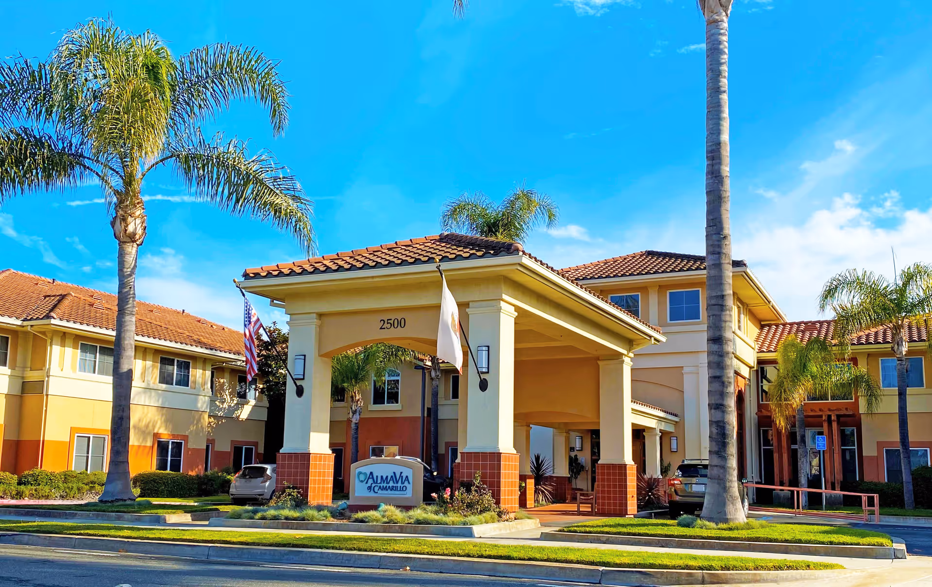 Exterior view of AlmaVia of Camarillo senior living facility with a covered entrance, palm trees, and clear blue sky.