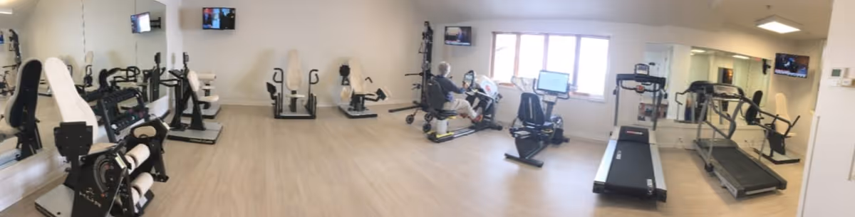 A senior living facility exercise room with various fitness equipment including recumbent bikes, treadmills, and strength training machines. A person is using one of the exercise machines near a window. The room has light wood flooring, white walls, and mirrors along one side. Several wall-mounted TVs are visible.