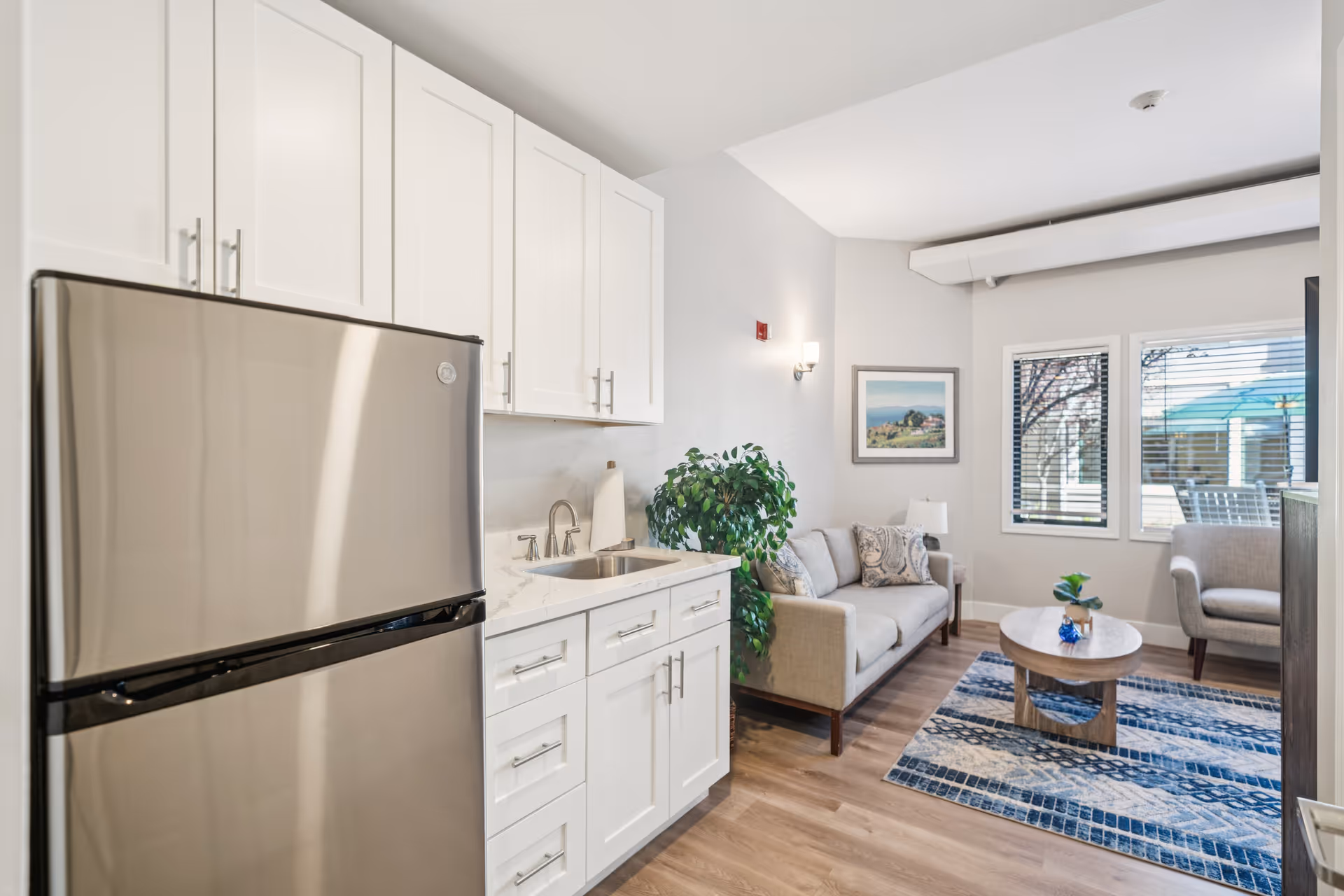 A bright and modern interior space featuring a small kitchenette with white cabinets, a stainless steel refrigerator, and a sink. Adjacent to the kitchenette is a cozy living area with a beige sofa, an armchair, a wooden coffee table on a blue patterned rug, a potted plant, and two windows with blinds letting in natural light.