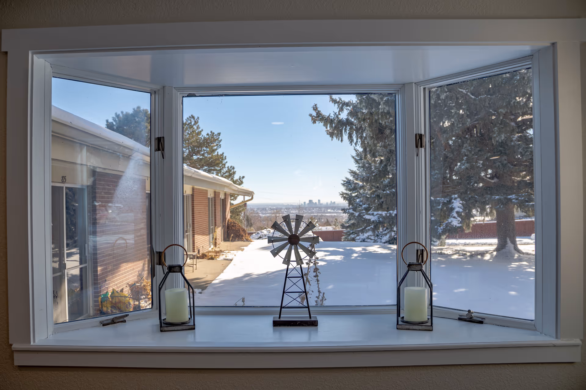 Bay window with two lantern candles and a small windmill sculpture looking out over a snowy yard and distant skyline.