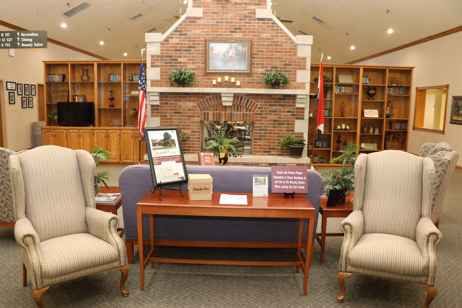 A cozy common area in a senior living facility featuring two striped armchairs facing a wooden table with informational signs and a donation box. Behind the table is a purple couch, and in the background, there is a large brick fireplace flanked by wooden bookshelves filled with books and decorative items. An American flag and another flag stand on either side of the fireplace. The room has a high ceiling with recessed lighting and a carpeted floor.