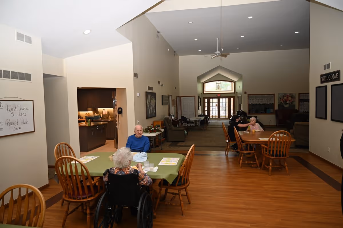 Interior of a senior living facility dining area with wooden floors and tables. Two elderly individuals are seated at a table in the foreground, one in a wheelchair and the other in a blue sweater. Another elderly person is seated at a table in the background being assisted by a caregiver. The room has high ceilings with recessed lighting and a ceiling fan. A kitchen area is visible through an open doorway on the left, and a welcome sign is on the right wall.
