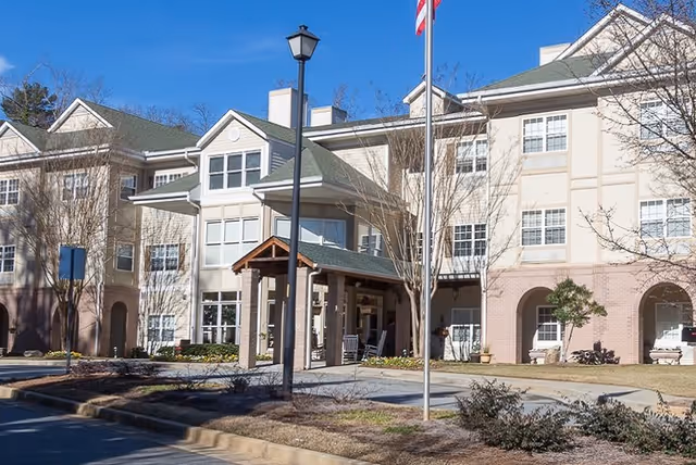 Exterior view of a multi-story senior living facility building with beige and brick facade, multiple windows, a covered entrance with rocking chairs, a flagpole with an American flag, and landscaped grounds with trees and shrubs.
