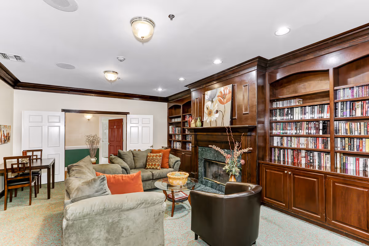 A cozy living room with green upholstered sofas and a brown leather armchair arranged around a glass coffee table. The room features a dark wood built-in bookshelf filled with books and a fireplace with a floral painting above it. There is a small table with chairs in the background near double white doors and a red door further back.