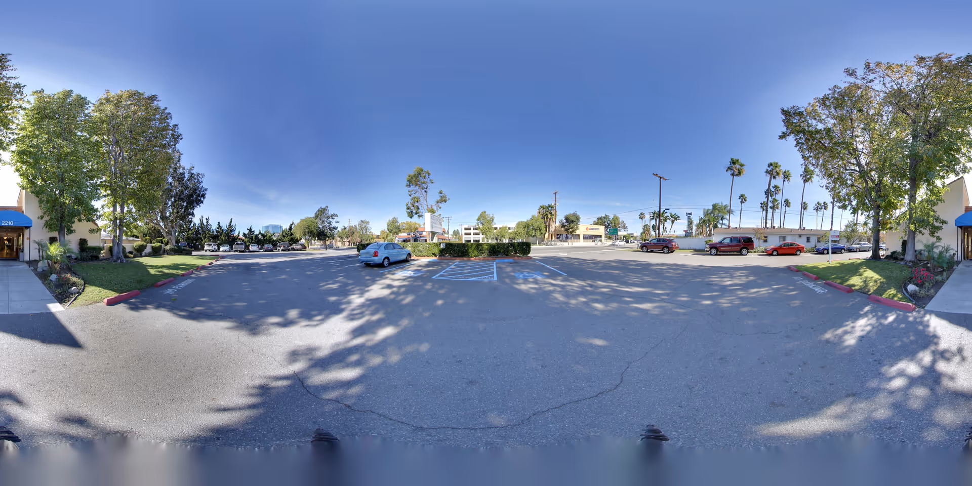 Wide view of a parking lot outside a building with trees and several parked cars under a clear blue sky. There are sidewalks and landscaped areas with grass and bushes on either side of the parking lot.