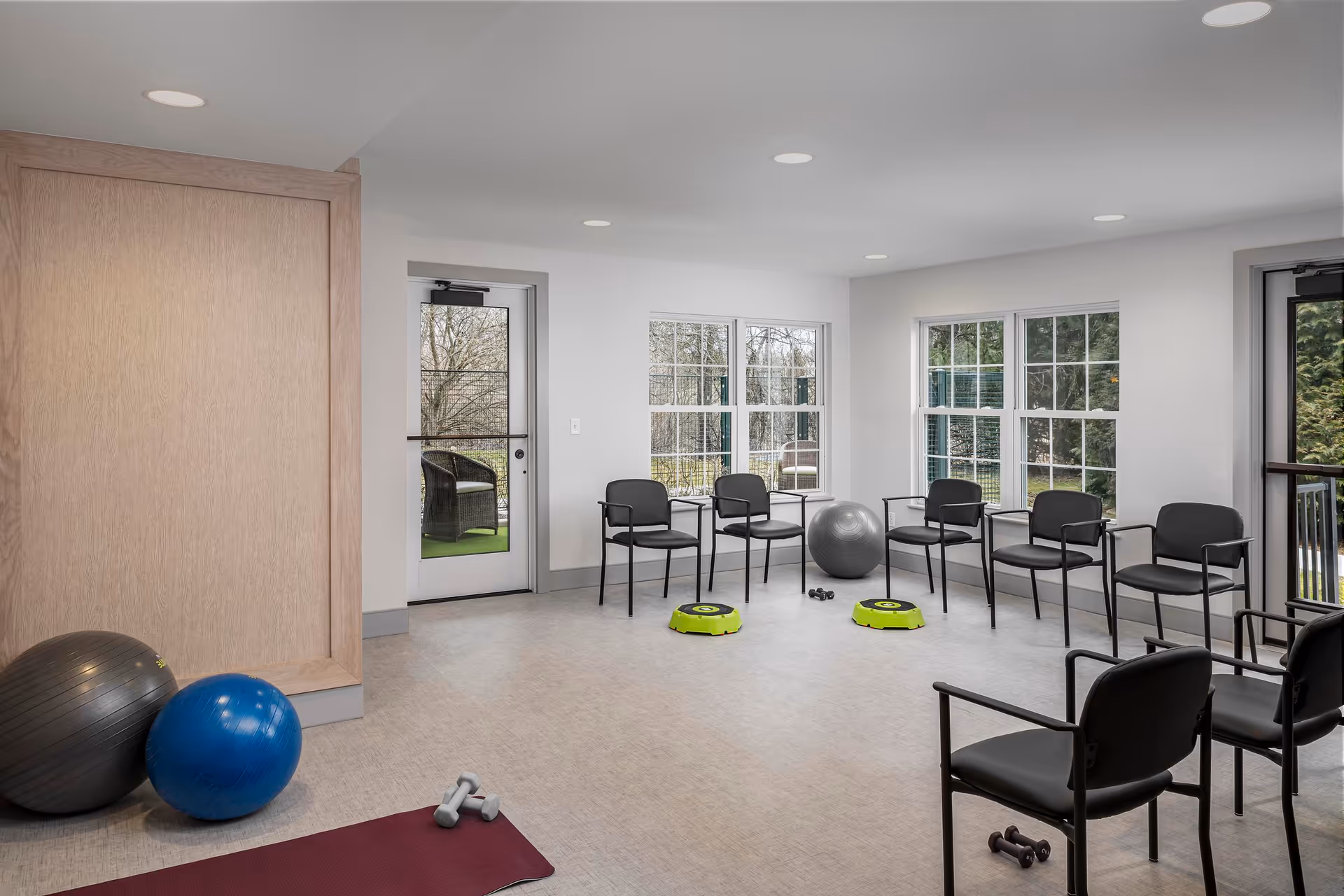 Bright exercise room with chairs arranged in a semicircle, exercise balls, mats, small dumbbells, and two green step platforms.