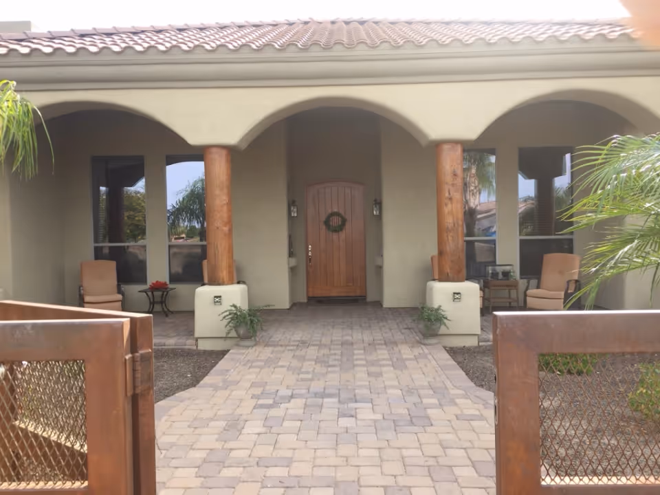 Paved walkway leading to the arched front porch of an assisted living building with wooden columns and a central wooden door.